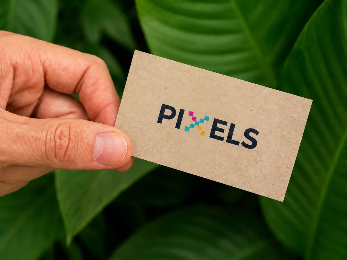 Mão segurando um cartão de visita PIXELS® em papel reciclado diante de folhagem verde